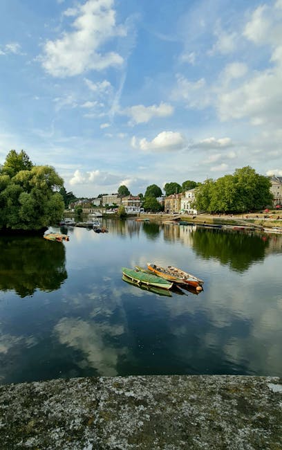 A tranquil river scene featuring a calm, reflective water surface with gentle ripples, bordered by lush green trees and shrubs on both sides. Several small boats, including a green and a beige boat, a